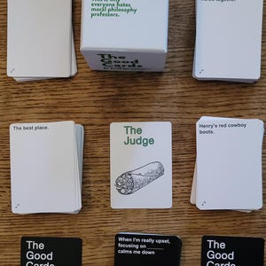 May include: A set of The Good Cards game with black and white cards featuring various prompts and phrases. The game box is white with green text and a green logo. The cards include prompts like "The best place", "The Judge", "Henry's red cowboy boots", "When I'm really upset, focusing on calms me down", and "Mashing your food holes together".