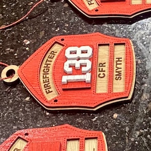 May include: Red and natural wood firefighter helmet ornaments. Each ornament features the word "FIREFIGHTER" in a curved design, a number, and the names of the firefighter and the fire department. The ornaments are made of layered wood and have a small hole for hanging.