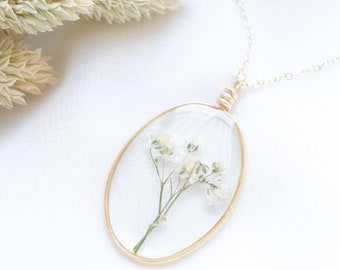Könnte beinhalten: Eine goldene Kettenkette mit einem klaren ovalen Anhänger, der eine gepresste weiße Blume enthält.
