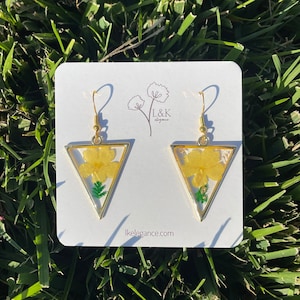 Puede incluir: Un par de pendientes triangulares de oro con flores amarillas prensadas y hojas verdes dentro de la resina. Los pendientes están en una tarjeta blanca con el texto "L&K elegance" y "lkelegance.com".