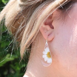 May include: A pair of teardrop-shaped earrings with a white flower pressed inside each clear resin drop.