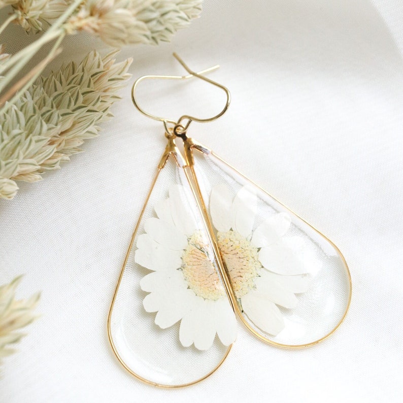 May include: A pair of gold teardrop-shaped earrings with a pressed white daisy flower inside each earring.