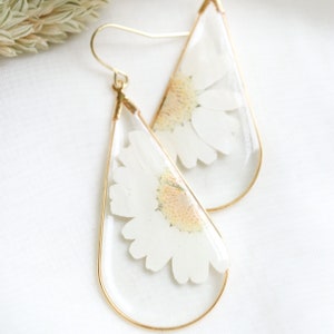 May include: A pair of gold teardrop-shaped earrings with a white pressed flower inside each earring.