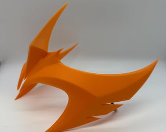May include: An orange, angular mask with sharp, pointed edges. The mask has a curved design with a central crest and side extensions. The material appears to be plastic or a similar lightweight material. The mask is likely for cosplay or costume use.
