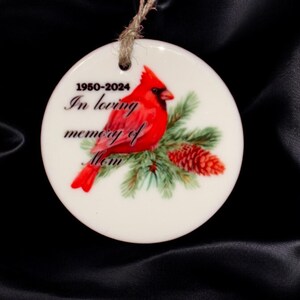 Può includere: Ciondolo in ceramica bianca con un cardinale rosso appollaiato su un ramo di pino. Il ciondolo ha il testo "1950-2024 In memoria affettuosa di Mamma".