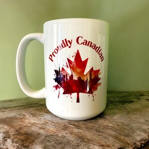 May include: White ceramic mug with a handle, featuring the text "Proudly Canadian" in red, and a red maple leaf design with a cityscape silhouette. The mug is on a wooden surface.