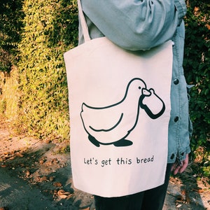 Könnte beinhalten: Eine weiße Canvas-Tragetasche mit einer schwarzen Illustration eines Entchens, das ein Brot trägt. Der Text "Let's get this bread" ist unter dem Entchen aufgedruckt.
