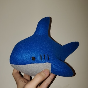 May include: A blue plush shark toy with a gray belly and black eyes. The shark has a stitched-on mouth with three rows of teeth.