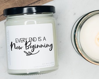 May include: A white soy wax candle in a clear glass jar with a black lid. The candle has a white label with the text "EVERY END IS A NEW BEGINNING" in black script font. The candle is on a white marble surface.