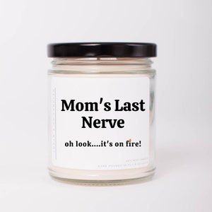 May include: A white candle jar with a black lid. The label reads "Mom's Last Nerve" and "oh look....it's on fire!"