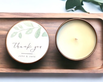 May include: A small, round candle with a white wax top and a green leafy design on the label. The label says "thank you" with the names "John & Jane" underneath. The candle is on a wooden tray.
