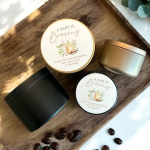 May include: Four small, round candles in black and gold tins, and two with labels that read "A Baby Is Brewing." The labels also include text: "THANK YOU FOR SHOWERING ANY WITH LOVE 07/04/2025." Coffee beans are scattered on a wooden tray.