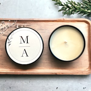 May include: A black candle with a white label that says "M/A" on a wooden tray. The candle is surrounded by greenery.