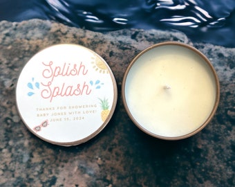 May include: A small, round, gold-colored tin with a white label that says "Splish Splash" in pink and blue lettering. The label also includes a yellow sun, blue water droplets, and a pineapple. The text "Thanks for showering Baby Jones with love! June 15, 2024" is printed on the label. The tin is open and a white candle is visible inside.
