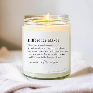May include: A glass jar candle with a white label that reads "Difference Maker /dif-er-uhns-mey-ker/noun A dedicated person who can make a big impact, even with just a small action or a few words. Someone who makes a difference in the lives of others. Also Known as: Ms. Amy SOY WAX CANDLE HAND POURED IN FL 8 OZ (226g)"