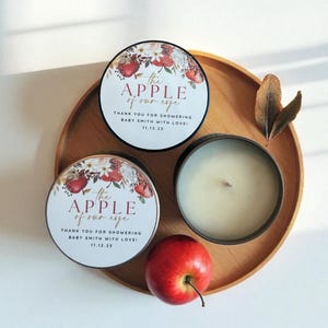 Peut inclure: Un plateau en bois pr&eacute;sente deux bo&icirc;tes &agrave; bougies rondes avec des motifs floraux et de pommes, et le texte "the APPLE of our eye". Une bo&icirc;te &agrave; bougie ouverte r&eacute;v&egrave;le une bougie blanche. Une pomme rouge est pos&eacute;e sur le plateau.