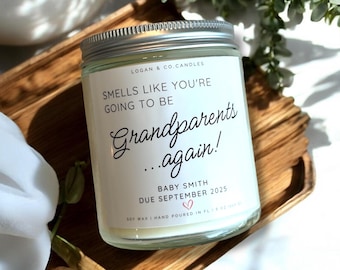May include: A clear glass candle jar with a silver lid, featuring a label that reads "Smells Like You're Going to Be Grandparents Again!" with the baby's name and due date. The candle sits on a wooden tray.