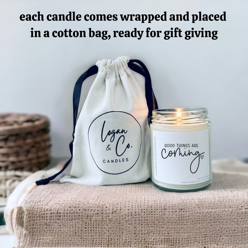 May include: A white cotton bag with a black drawstring and a black logo that says "Logan & Co. Candles" is next to a clear glass candle with a white label that says "Good things are coming" with a heart symbol.