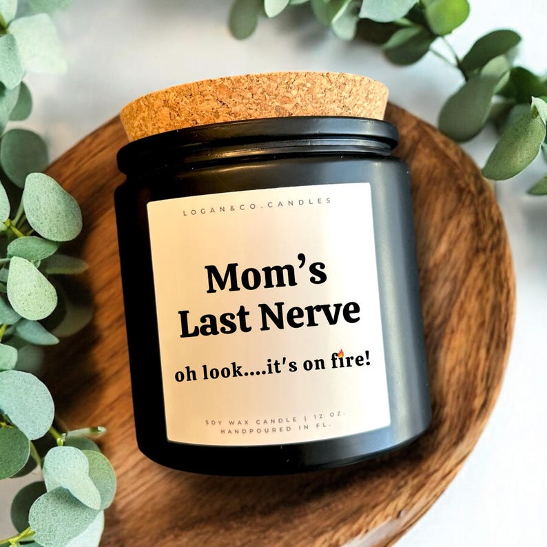 May include: Black soy wax candle with a cork lid. The label reads "Mom's Last Nerve oh look....it's on fire!" and "LOGAN & CO. CANDLES". The candle is 12 oz and hand-poured in Florida.