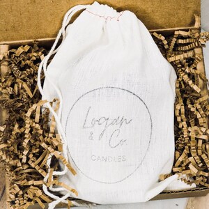 K&ouml;nnte beinhalten: Ein wei&szlig;er Kordelzugbeutel mit dem Text "Logan & Co. Candles" in einer Pappschachtel, die mit braunem Krepppapier gef&uuml;llt ist. Die Schachtel ist ge&ouml;ffnet und zeigt den Inhalt.
