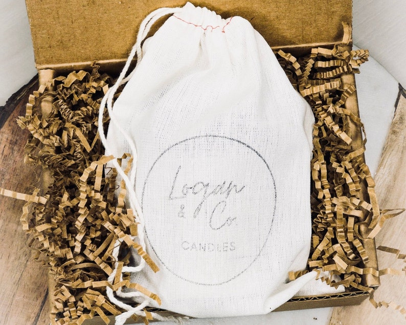 K&ouml;nnte beinhalten: Ein wei&szlig;er Kordelzugbeutel mit dem Text "Logan & Co. Candles" in einer Pappschachtel, die mit braunem Krepppapier gef&uuml;llt ist. Die Schachtel ist ge&ouml;ffnet und zeigt den Inhalt.