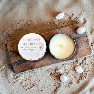 May include: Two round, metal-rimmed candles on a wooden board, one with a lid featuring text and floral designs. The text reads "a little pearl is on her way!" and "Thank you for showering Emily with love! May 9, 2026." Scattered around are pearls and seashells.