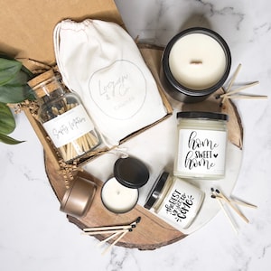 May include: A gift set for home fragrance lovers featuring three candles, a jar of matches, and a muslin bag. The candles are in glass jars with black lids and labels that say "Home Sweet Home" and "The Best is Yet to Come". The matches are in a clear glass jar with a cork lid and a label that says "Safety Matches". The muslin bag is white with a logo that says "Logan & Co Candles".
