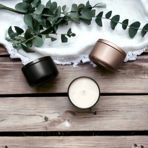 Peut inclure: Trois petites bougies rondes avec couvercles noirs et or rose, et une bougie ouverte avec un int&eacute;rieur en cire blanche. Les bougies sont sur une surface en bois rustique, avec une branche d'eucalyptus vert et un napperon en dentelle blanche.