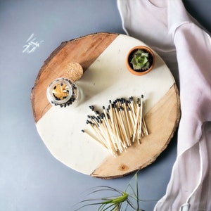May include: A wooden tray with a white marble insert. The tray has a small glass jar filled with matches and a small terracotta pot with a green succulent plant. The tray is decorated with a scattering of black-tipped matches.
