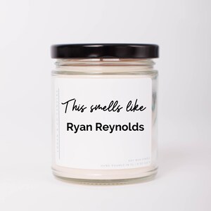 May include: A white candle in a clear glass jar with a black lid. The label on the jar reads "This smells like Ryan Reynolds".