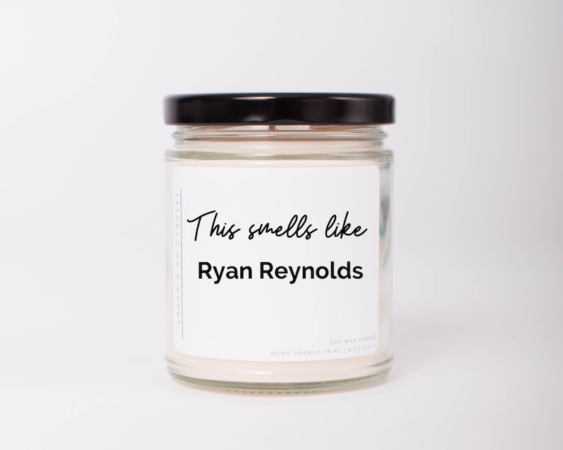 May include: A white candle in a clear glass jar with a black lid. The label on the jar reads "This smells like Ryan Reynolds".