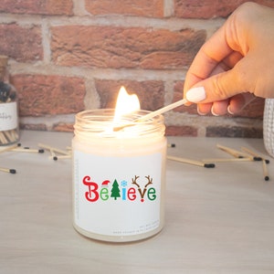 K&ouml;nnte beinhalten: Eine brennende Sojawachskerze in einem Glas mit dem Wort "BELIEVE" in Rot, Gr&uuml;n und Blau, mit weihnachtlichen Symbolen. Eine Hand z&uuml;ndet die Kerze mit einem Streichholz an. Ein Glas mit Streichh&ouml;lzern ist im Hintergrund.