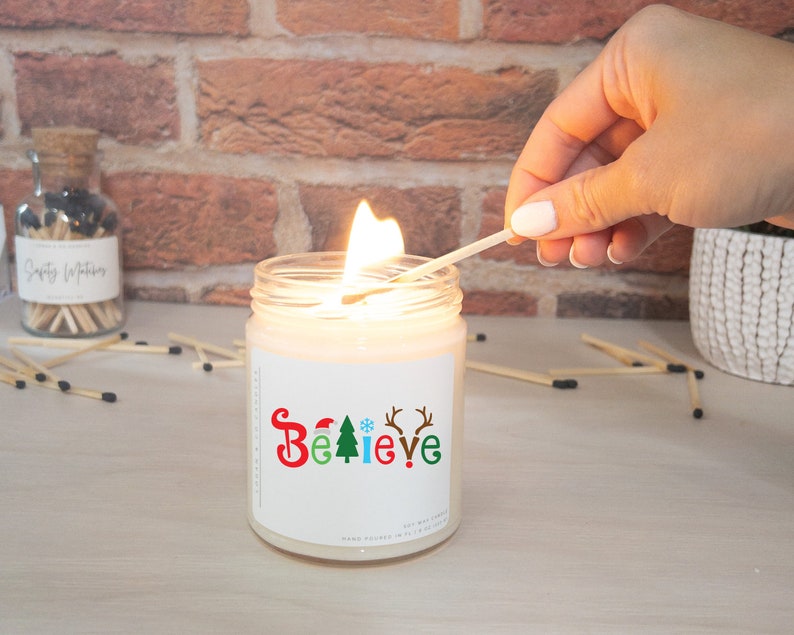 K&ouml;nnte beinhalten: Eine brennende Sojawachskerze in einem Glas mit dem Wort "BELIEVE" in Rot, Gr&uuml;n und Blau, mit weihnachtlichen Symbolen. Eine Hand z&uuml;ndet die Kerze mit einem Streichholz an. Ein Glas mit Streichh&ouml;lzern ist im Hintergrund.