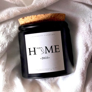 May include: Black glass candle jar with a cork lid. The label is white with black text that reads "LOGAN & CO. CANDLES" and "HOME" with an outline of the state of Florida. The text "2023" is centered below the word "HOME". The bottom of the label reads "SOY WAX CANDLE | 13 OZ HAND POURED IN FL."