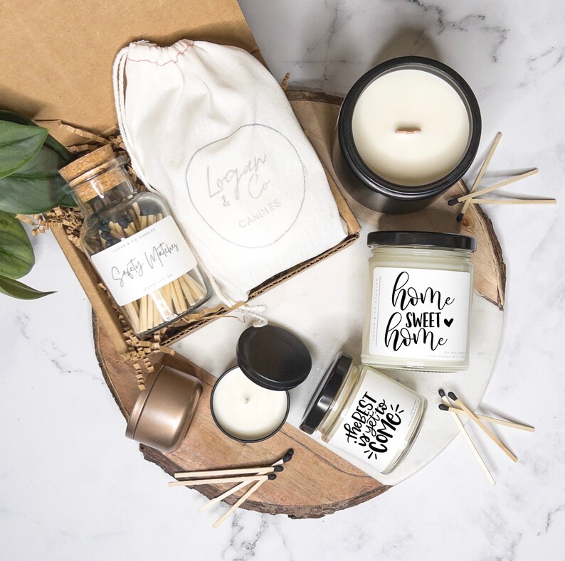 May include: A gift set for home fragrance lovers featuring three candles, a jar of matches, and a muslin bag. The candles are in glass jars with black lids and labels that say "Home Sweet Home" and "The Best is Yet to Come". The matches are in a clear glass jar with a cork lid and a label that says "Safety Matches". The muslin bag is white with a logo that says "Logan & Co Candles".