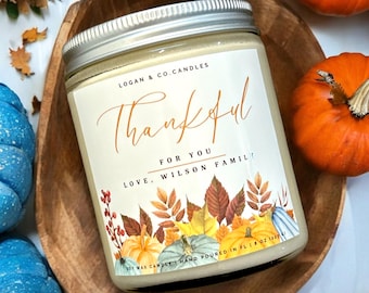 May include: A white candle with a label that says "Thankful for you Love, Wilson Family". The label has a fall-themed design with pumpkins, leaves, and berries. The candle is in a glass jar with a silver lid. The candle is sitting on a wooden surface.
