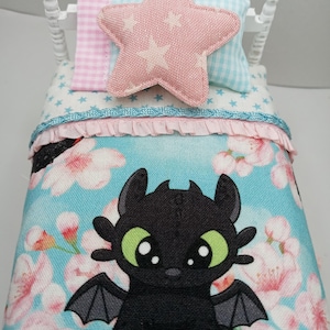May include: A miniature white bed with a blue floral patterned comforter featuring a black dragon character. The bed has a pink star-shaped pillow and two smaller pillows. The bed frame is white.