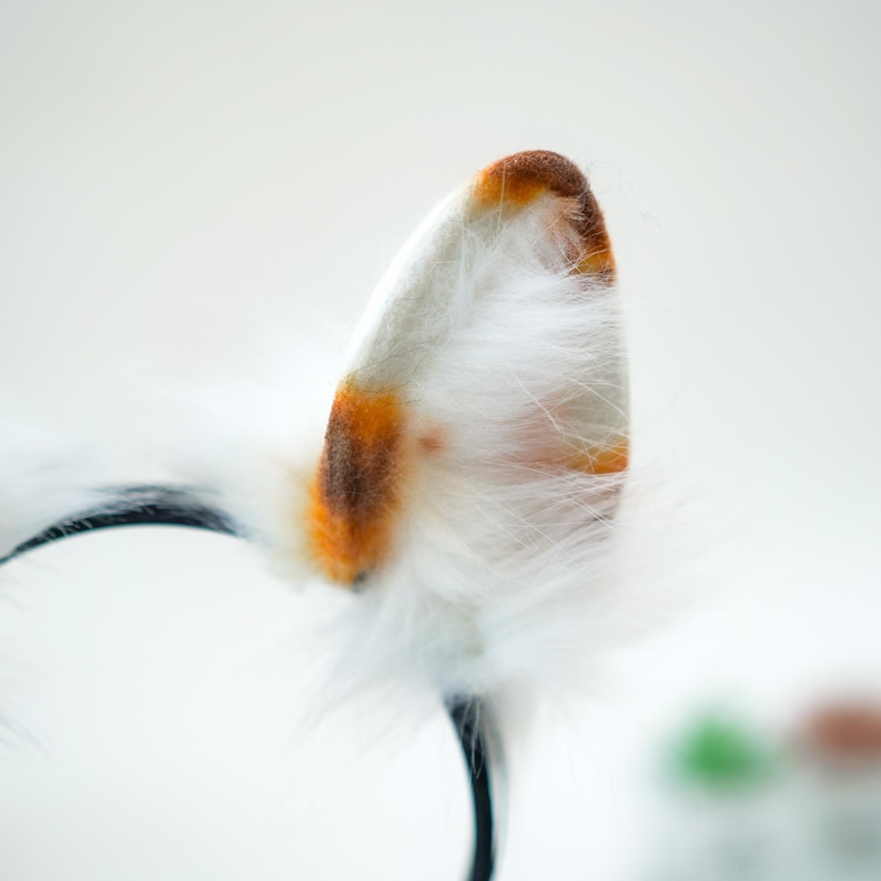 May include: A pair of cat ears on a black headband. The ears are white with orange and brown accents and fluffy white fur. The headband is black.
