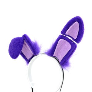 May include: A headband featuring two plush purple rabbit ears with light purple inner panels. The left ear is folded downwards, while the right ear is upright and articulated with a visible hinge, showcasing a soft, fuzzy texture throughout.