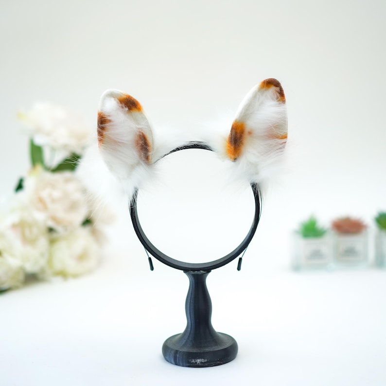 May include: White and brown faux fur cat ears headband. The ears have brown spots and are attached to a black headband. The headband is displayed on a black stand against a white background.