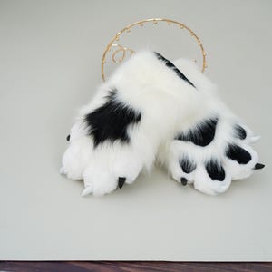 May include: A pair of plush, white and black paw-shaped gloves. The gloves have a fluffy texture with black spots and black claws. The gloves are designed to resemble animal paws.