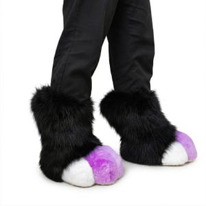 Puede incluir: Pies de disfraz de piel sintética negra con almohadillas blancas y moradas. Las botas de piel sintética se usan con pantalones negros. Las botas están diseñadas para parecerse a las patas de los animales.