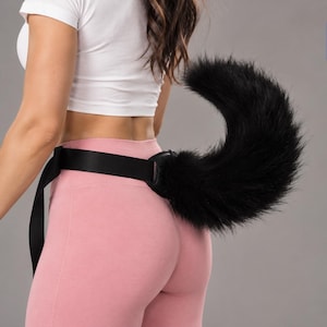 May include: A black faux fur animal tail accessory attached to a wide black fabric belt. The tail is long, thick, and curved, featuring dense, shaggy black synthetic fibers that extend from a reinforced black base connected to the adjustable strap.