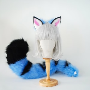May include: A set of faux fur animal ears and a long, striped tail featuring vibrant blue and black fur. The ears have pink inner detailing and are attached to a headband. The set is displayed with a short, straight, silver-white wig.