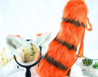 May include: Orange and black striped faux fur tail with a white fluffy tip and a matching headband with white fluffy ears. The tail is tied with a bright orange ribbon.