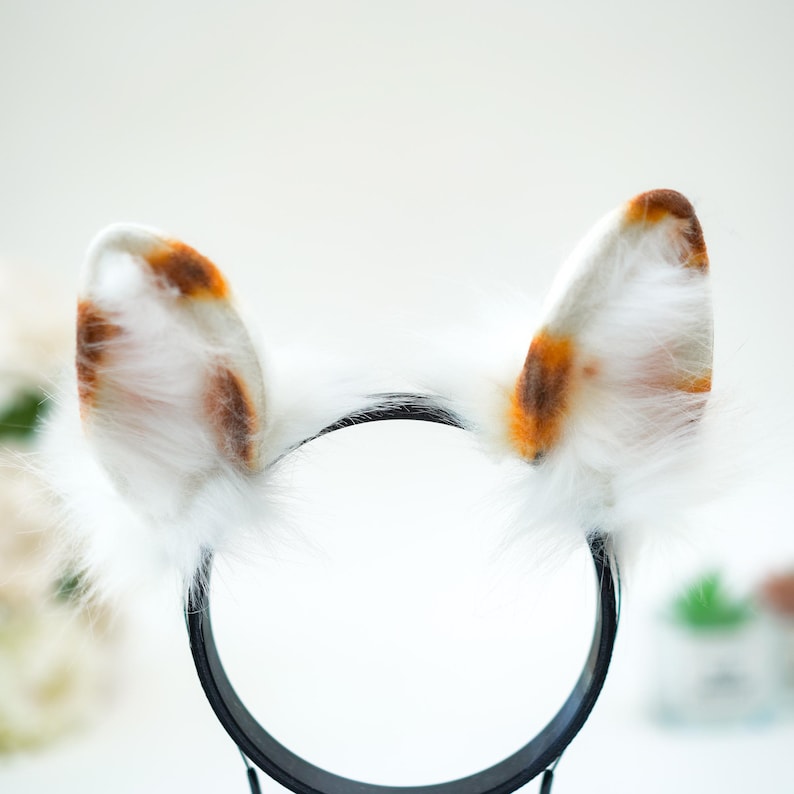 May include: A pair of cat ear headbands. The ears are white with brown and orange spots, and are made of fluffy faux fur. The headband is black and has a simple, rounded design. The ears are upright and symmetrical, giving a playful look.
