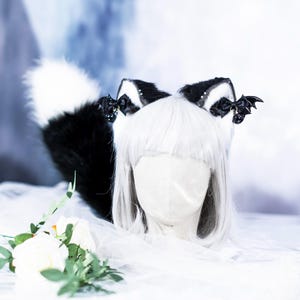 Peut inclure: Un serre-tête avec des oreilles de chat en fausse fourrure noire et blanche, accompagné d'une queue assortie. Les oreilles sont ornées de nœuds noirs et d'un charm en forme de chauve-souris. La queue est touffue. Un accessoire unique pour le cosplay.