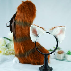 Pode incluir: Uma faixa de cabeça com orelhas de gato de pele sintética branca com uma faixa de cabeça preta. A faixa de cabeça está em um suporte na frente de uma cauda vermelha e marrom fofa.
