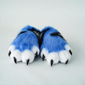 May include: A pair of furry blue and white paw-shaped slippers. The slippers have black claws and black stripes on the top. The slippers are designed to resemble animal paws.