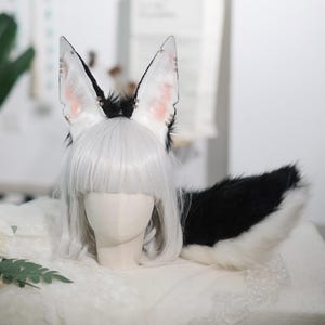 Może przedstawiać: Zestaw cosplay z uszami lisa i ogonem. Uszy są białe z czarnymi końcówkami i różowymi detalami wewnętrznymi, ozdobione małymi ozdobami. Ogon jest czarny z białą końcówką. Zestaw jest prezentowany na głowie manekina z białą peruką.